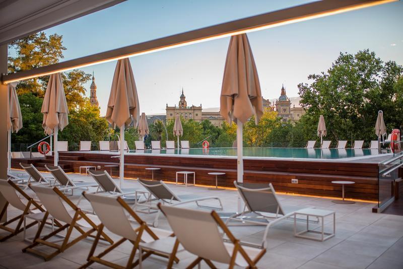 Outdoor terrace with lounge chairs, pool and umbrellas overlooking city rooftops.